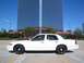 1998 Ford Crown Victoria - A white 1998 Ford Crown Victoria sedan is seen from the side, parked in front of a large, modern glass building under a clear blue sky.
