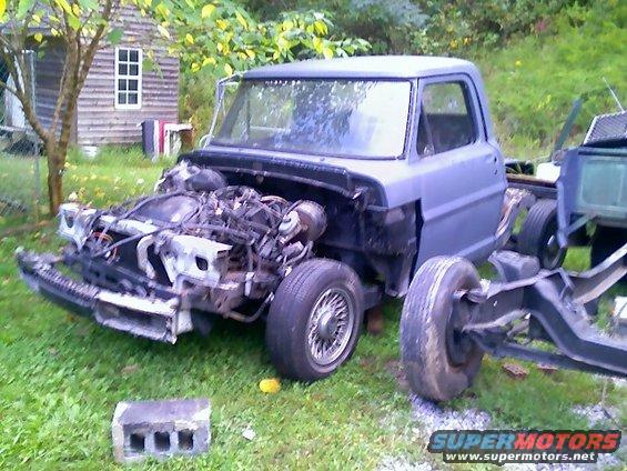 Disassembled Ford truck chassis with engine exposed, missing body panels, and one wheel detached