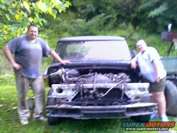Frontal view of a dark-colored truck with its hood open and engine exposed, showing significant disrepair and missing parts