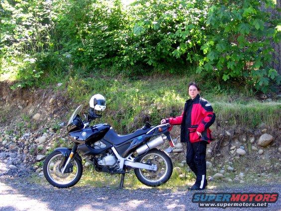 Person in riding gear stands next to a black dual-sport motorcycle with a white helmet on its handlebars