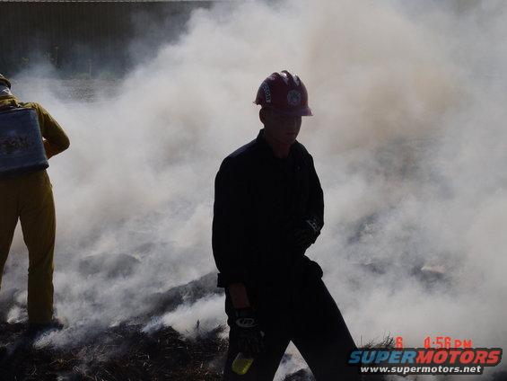 Two firefighters in protective gear stand in smoke and low flames