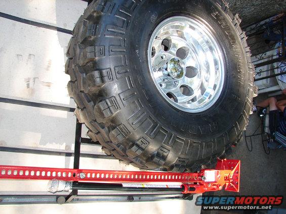 Off-road tire mounted on a chrome rim, with a red Hi-Lift jack positioned below it