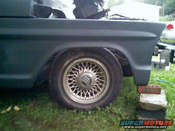 Close-up of a wire-spoke wheel with a Mercury logo on a dark vehicle. The vehicle is on bricks