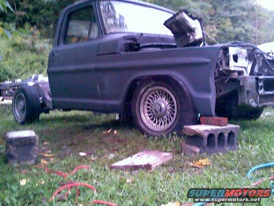 Dark gray Ford F-series truck body on blocks, missing front end and hood, with aftermarket wheels