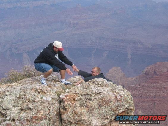 Man in black hoodie and blue shorts reaching down to help another man clinging to a rocky cliff edge in a canyon