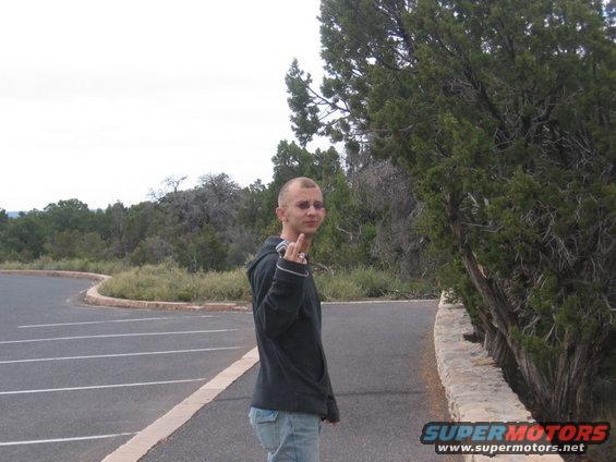 Man with shaved head and beard, wearing a dark hoodie and jeans, gives the middle finger while standing on a paved road next to trees