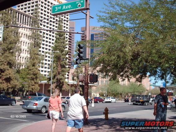 Street corner with traffic light, street signs for 3rd Ave, and people walking. Cars are visible on the road