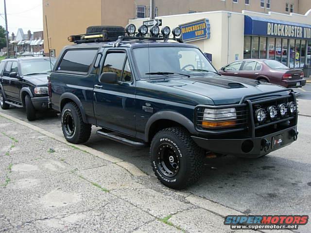 Dark green Ford Bronco with roof rack and brush guard parked next to a black SUV. Blockbuster Video in background