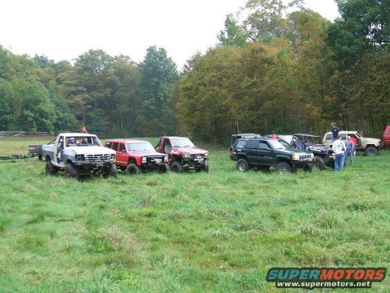 Several modified off-road vehicles parked in a grassy field with trees in the background