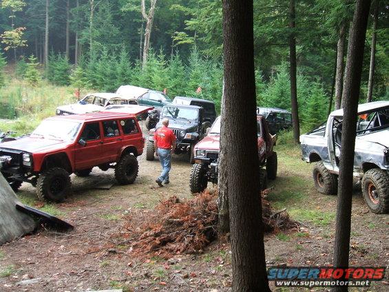 Several modified off-road vehicles parked in a forest clearing. One person walks between them