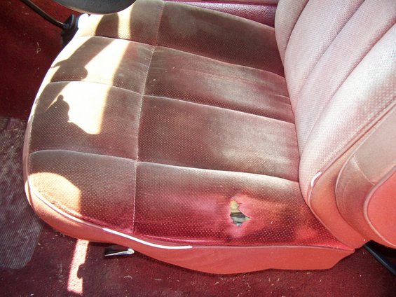 Close-up of a worn car seat with a large tear on the right side. The upholstery is red velour