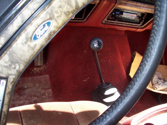 Interior shot of a 1985 Ford Bronco, showing the steering wheel with a Ford logo, the gear shifter, and the dashboard with a radio