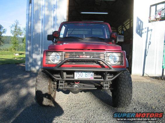 Front view of a red 1987 Suzuki Samurai with a custom bumper and off-road tires parked in front of a metal building