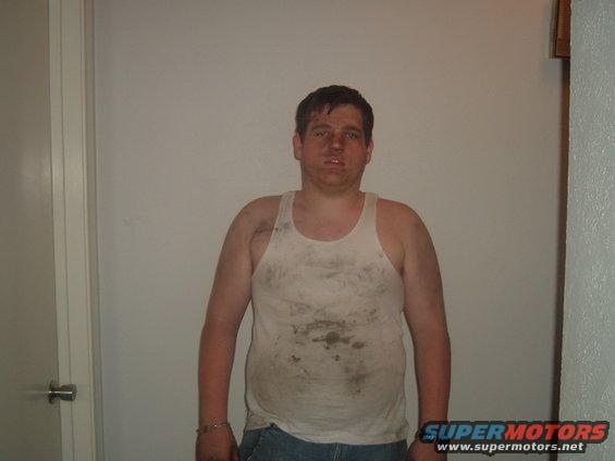 Man in dirty white tank top and jeans, face smudged with dirt, stands indoors