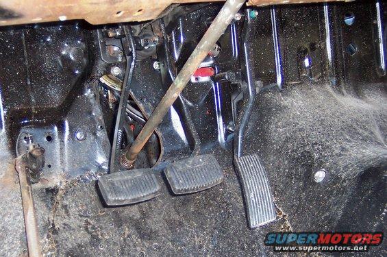 Close-up of vehicle floor with clutch, brake, and accelerator pedals visible, along with linkage and wiring