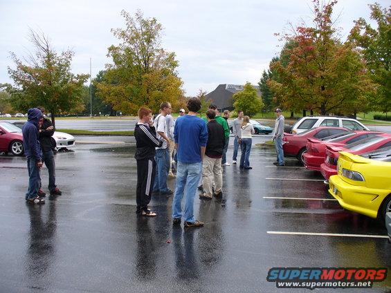 group of people standing around in a wet parking lot with several cars parked