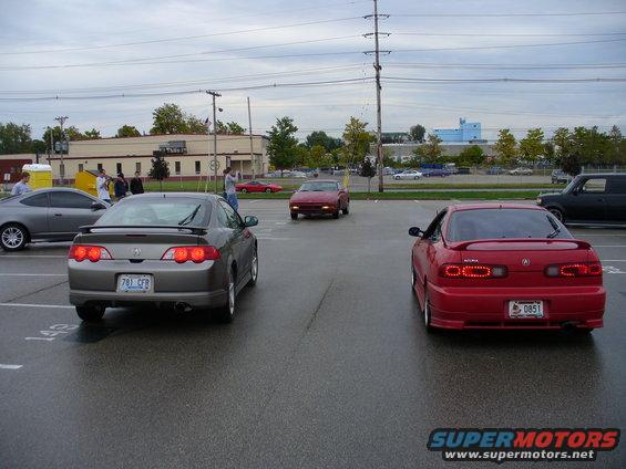 Two Acura Integra coupes, one gray and one red, parked in a lot. A red car is driving away in the background