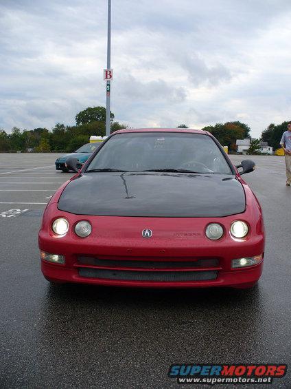 Front view of a red Acura Integra with a black hood parked in a lot. Another car is visible in the background
