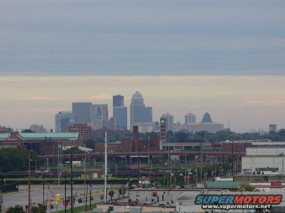 Cityscape with skyscrapers, a clock tower, and industrial buildings under a cloudy sky. Text 'SUPERMOTORS' is visible