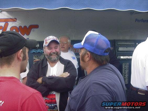 Three men in hats talking outdoors, with a blue awning and trailer in the background