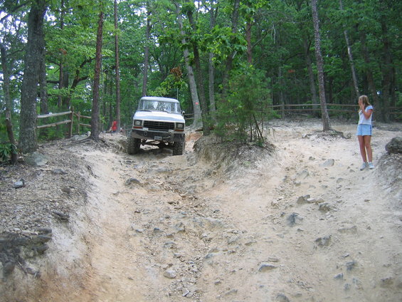 White off-road vehicle climbing a rocky dirt trail in a wooded area. A young woman stands to the right