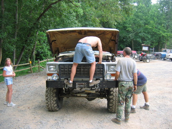 Truck with hood open, front bumper, winch, and large tires. Three people are working on it, one inside the engine bay. A young girl stands nearby