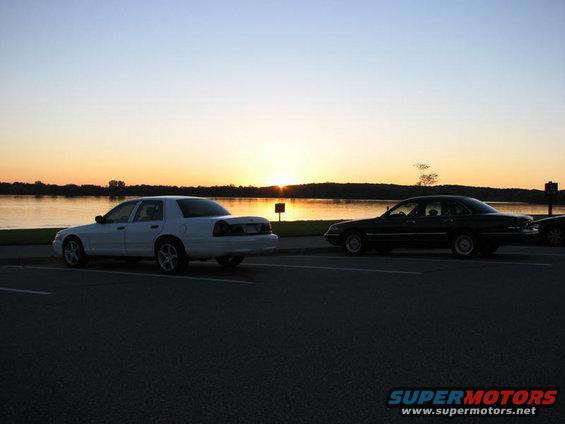 White 2003 Ford Crown Victoria and a dark sedan parked side-by-side facing a lake at sunset