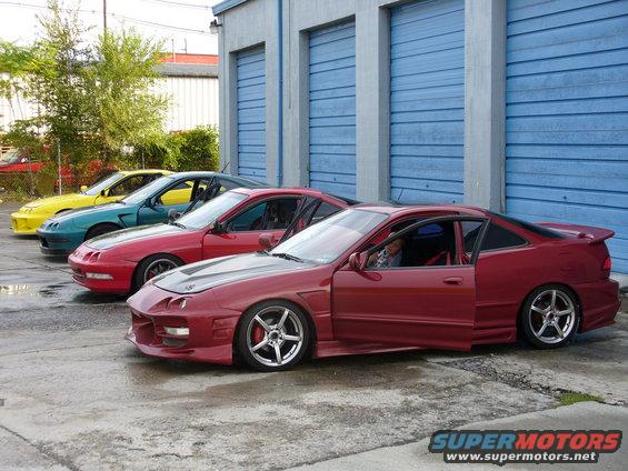 Four coupes parked in a row, three red, one teal, one yellow. The front red car has its doors open