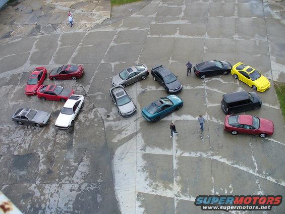 Several cars parked in a lot, including a red coupe, a white coupe, a silver sedan, a teal coupe, a black SUV, a yellow sedan, and a red hatchback