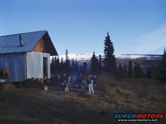 Cabin with a metal roof, people gathered around a campfire in a grassy, wooded area with snow-capped mountains in the background