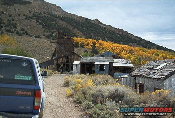 Blue pickup truck rear view, with "Frontier SE V6" visible on the tailgate, parked on a dirt road near abandoned buildings