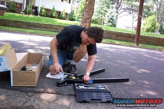 Man kneeling on asphalt, assembling a tow hitch with tools and parts from a box