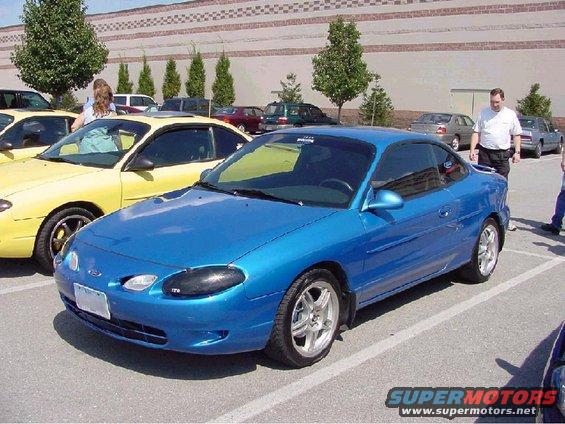 Blue coupe parked in a lot next to a yellow car. People are standing around