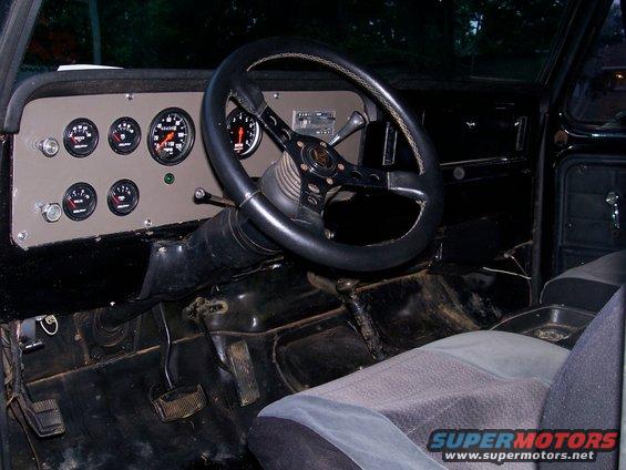 Interior shot of a vehicle dashboard with multiple gauges, a steering wheel, and part of a seat. A watermark reads SUPERMOTORS.NET