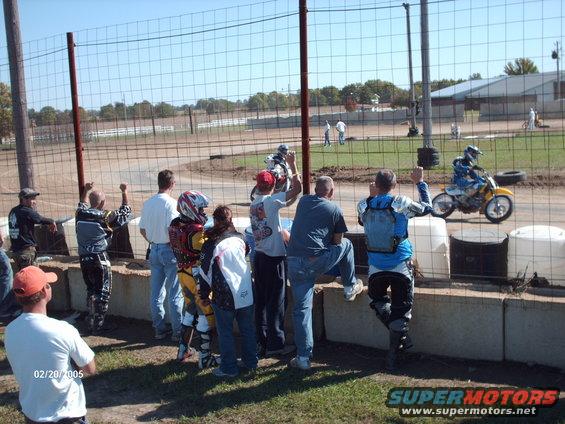 People watch a dirt bike race from behind a fence. One rider is on the track