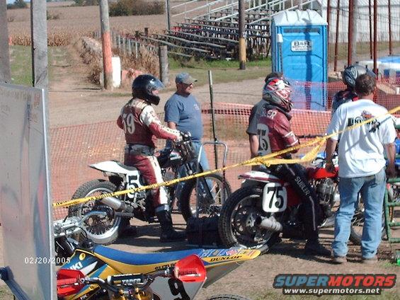 Motorcycles and racers at a dirt track with bleachers and a portable toilet in the background
