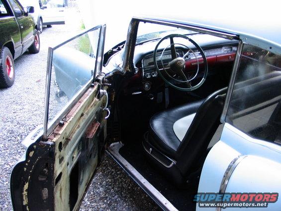 Interior of a light blue 1954 Cadillac with black and white seats and a green steering wheel. The driver's door is open, showing rust and missing panels