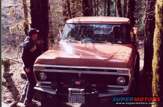 Front view of a red pickup truck parked in a forest. A person stands next to it
