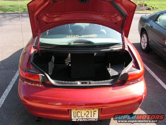 Trunk open on red sedan, showing subwoofer box and other items. Another car is parked next to it