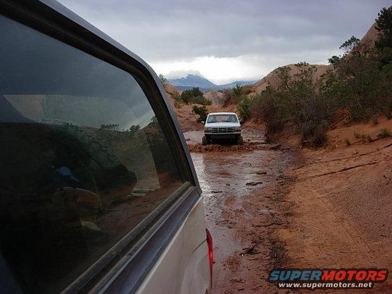 White pickup truck driving through a muddy trail in a desert landscape. Another vehicle is visible in the foreground