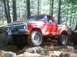 1976 Ford F-100 - A red Ford pickup truck navigates rough terrain in a wooded area, showing its lifted suspension and off-road tires.