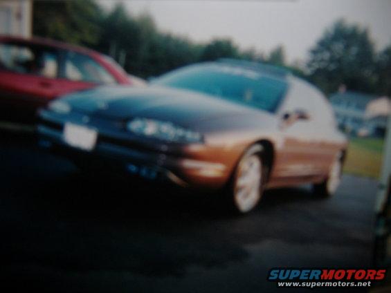Brown coupe parked next to a red car, with trees and houses in the background