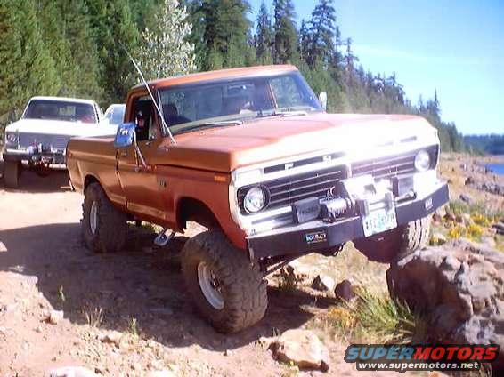 Orange pickup truck with large tires climbing over rocks, another white pickup truck behind it