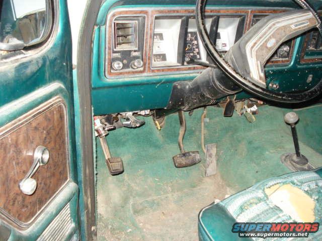 Green interior of a 1978 Ford Bronco with wood-grain door panel, pedals, steering wheel, and dashboard visible