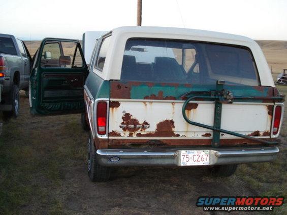 Rear view of a rusty green and white Ford Bronco with its driver's door open, parked next to a pickup truck