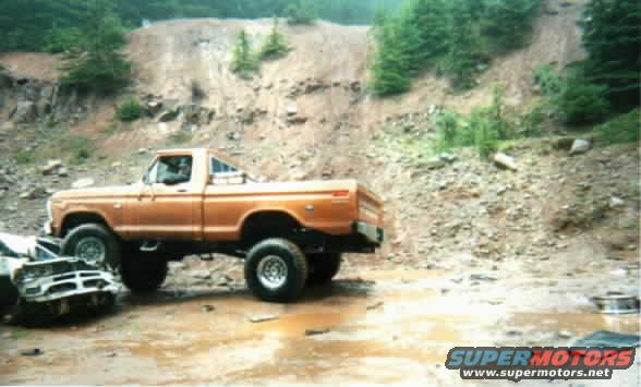 Orange pickup truck parked on muddy ground next to a wrecked car, with a dirt hill in the background