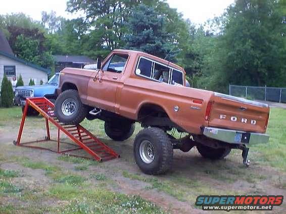 Orange Ford F-100 pickup truck on a ramp, with a blue truck in the background