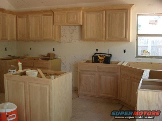 Unfinished kitchen cabinets with light wood doors and drawers, a window with a yellow sticker, and a black DeWalt tool case