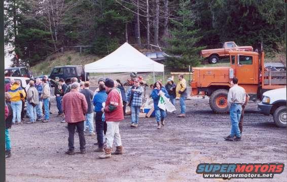 Group of people gathered around an orange truck and other vehicles in a dirt lot with trees in the background