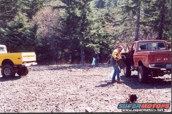Red Ford pickup truck parked on gravel with door open, yellow pickup truck in background